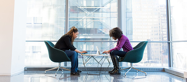 two-women-on-laptops-in-front-of-large-glass-window